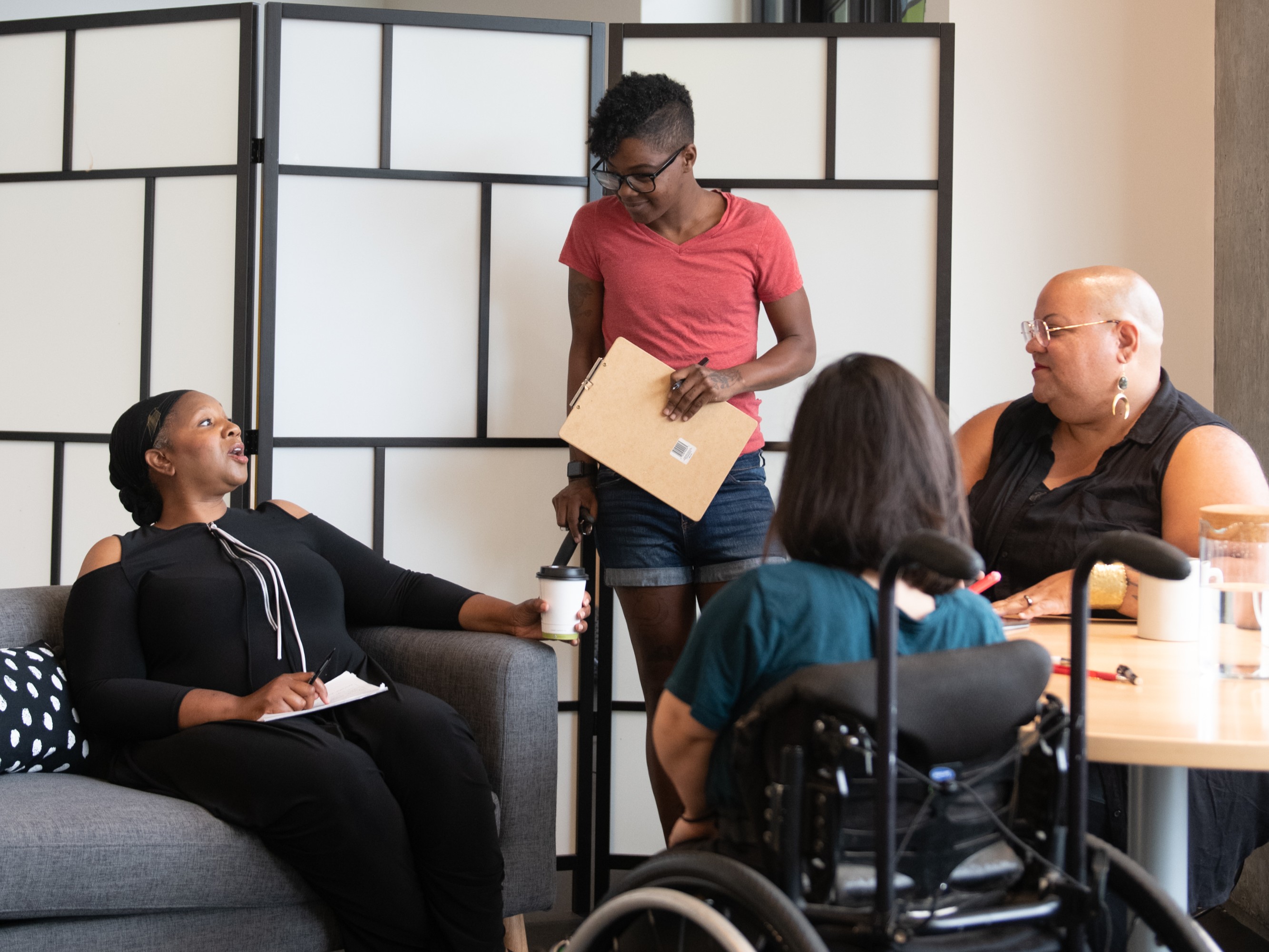 four disabled people of colour gather at a meeting. one sits on the couch, one stands with a cane, another sits at a table, and another sits in a wheelchair and takes notes.