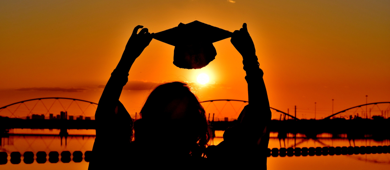 graduate holding cap in the air against a sunset background.