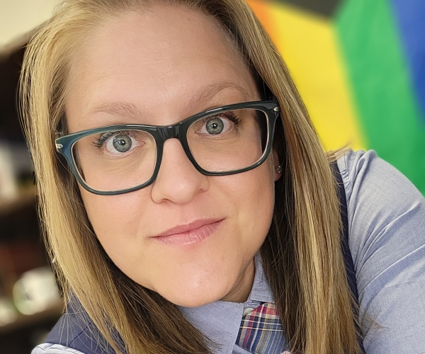 Ashlyne smiles at the camera, wearing a tie, vest, and collared shirt with the pride flag in the background